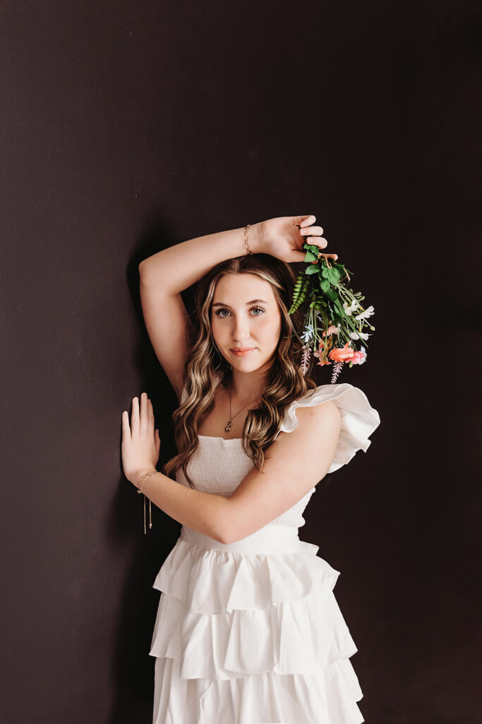 Teen girl standing against black wall in white dress, arms up and flowers at Reflection Images by Tracy Rowell in Florence SC