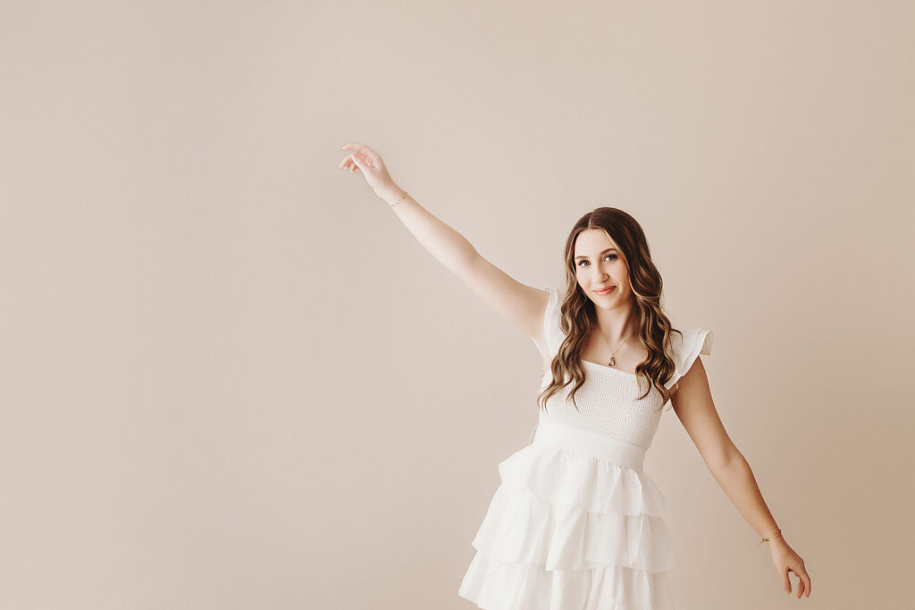 Teen girl standing against tan wall in white dress with arm up at Reflection Images by Tracy Rowell in Florence SC