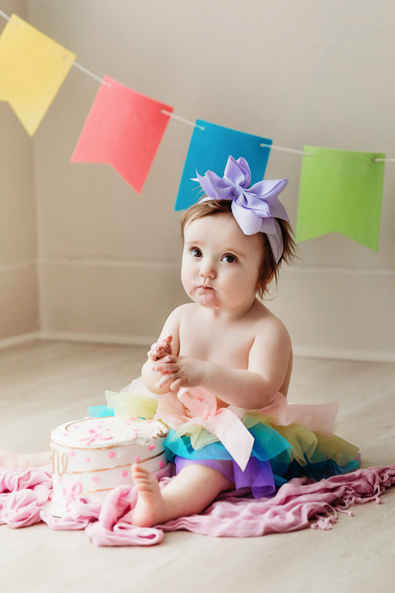 Sullivan joyfully smashing cake with frosting-covered toes and rainbow skirt in front of colorful birthday banners at Reflection Images by Tracy Rowell in Florence SC.