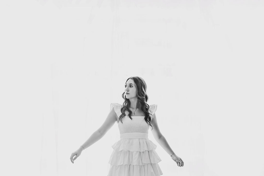 black and white of Teen girl standing against wall in white dress with arms out at Reflection Images by Tracy Rowell in Florence SC