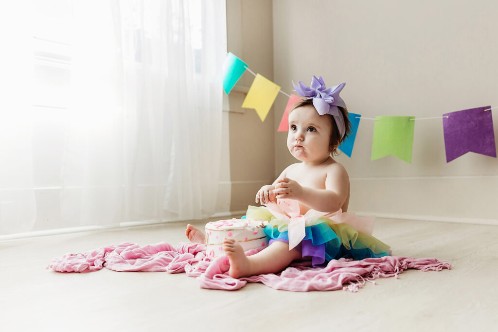 Sullivan digging into her cake during a birthday smash, wearing a rainbow tutu and lavender bow with colorful banners behind her at Reflection Images by Tracy Rowell in Florence SC.
