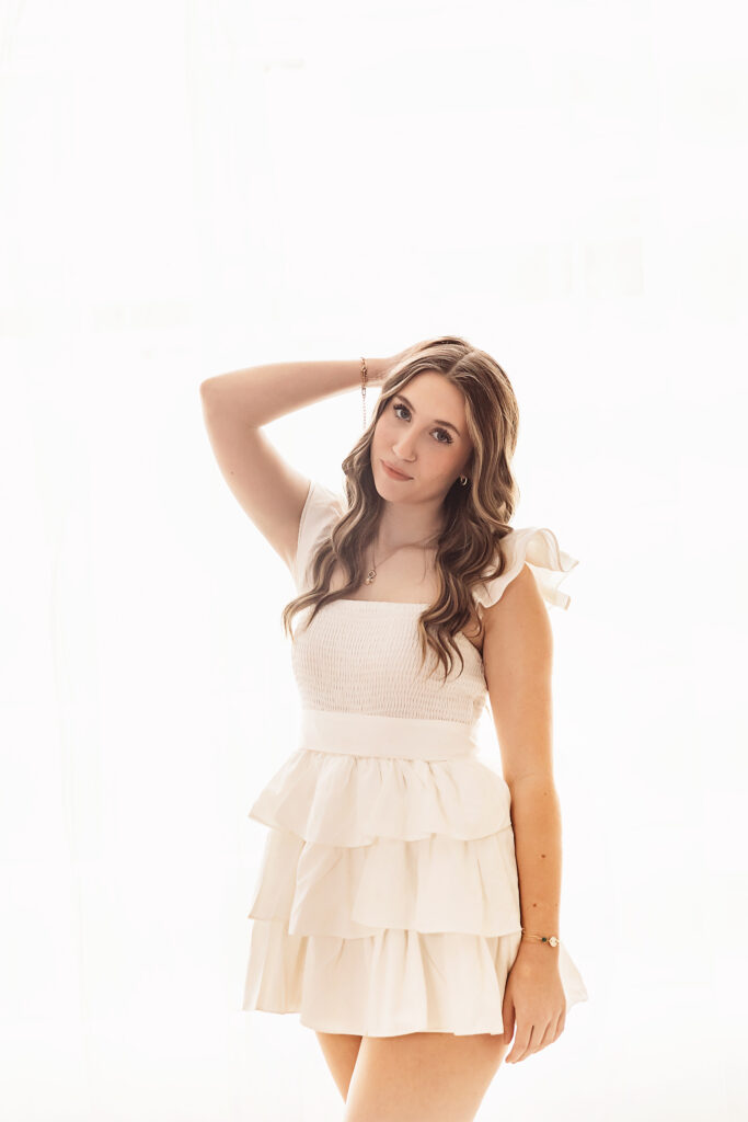 Teen girl standing against window wall in white dress with arm up smiling at Reflection Images by Tracy Rowell in Florence SC