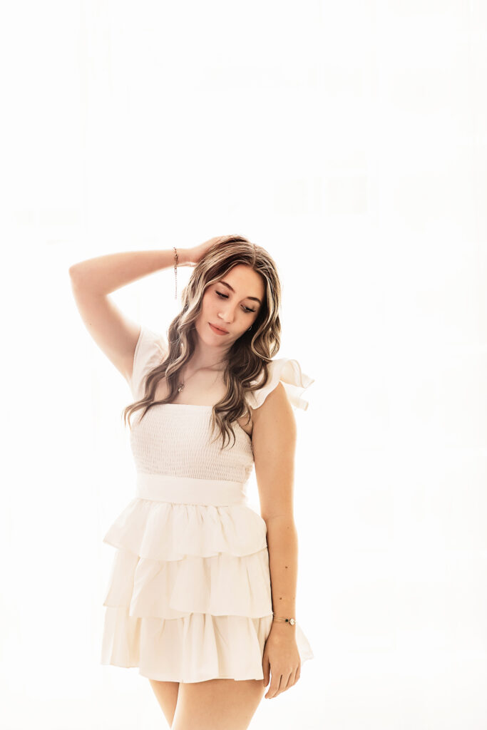 Teen girl standing against window wall in white dress with arm up looking down at Reflection Images by Tracy Rowell in Florence SC