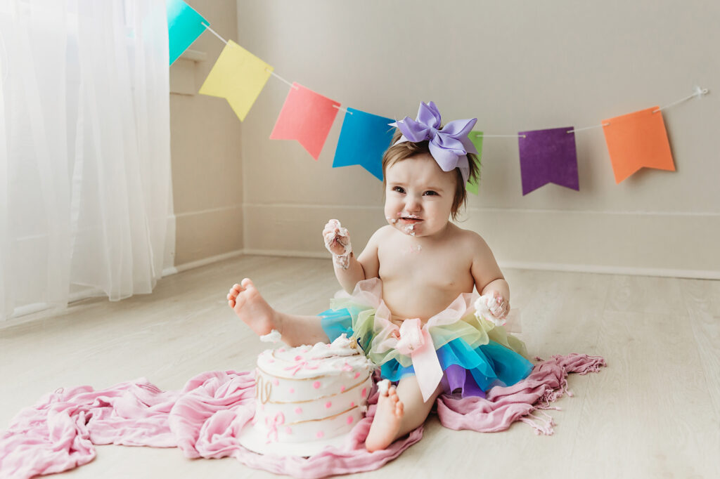 Front view of Sullivan enjoying her cake smash session in a rainbow skirt with soft frosting-covered hands at Reflection Images by Tracy Rowell in Florence SC.