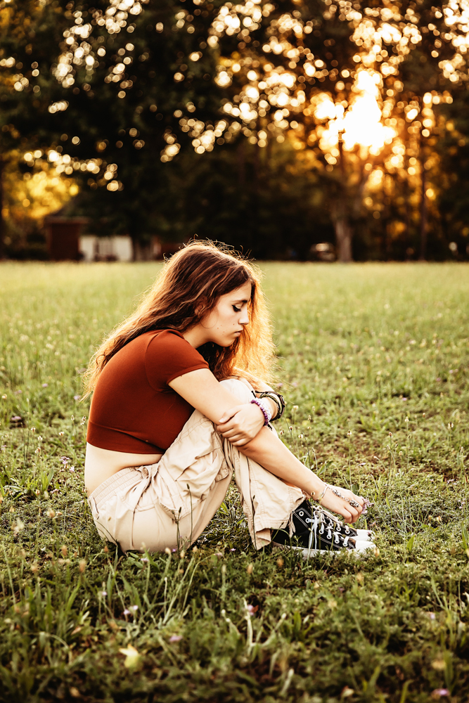 Eve sits curled up in the grass at sunset, wearing a rust-colored top and khaki pants, Converse sneakers on her feet.
