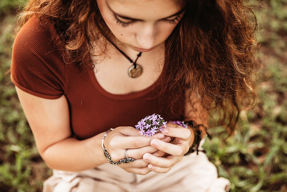 Close-up of Eve holding a small bunch of purple flowers, her necklace and rust top visible as she gazes down.