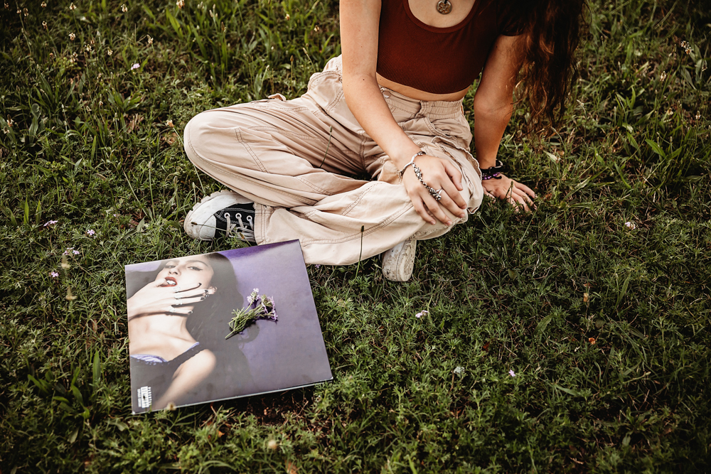 Artistic shot of Eves legs crossed in the grass beside a vinyl record cover, with purple flowers resting on top of it.