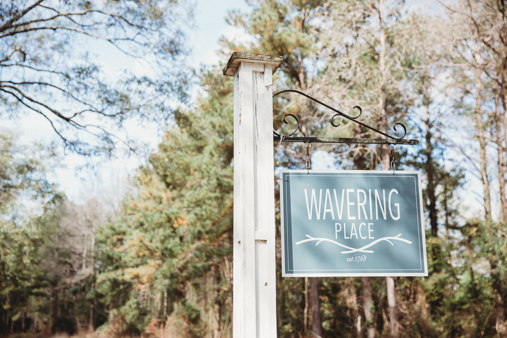 The Wavering Place venue sign hanging on a white post, surrounded by tall pine trees under soft morning light in Eastover, South Carolina.