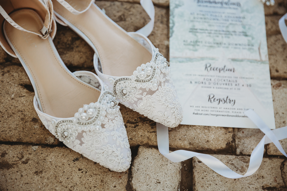 Delicate lace bridal shoes adorned with pearls placed beside invitation suite on textured brick surface at Wavering Place.
