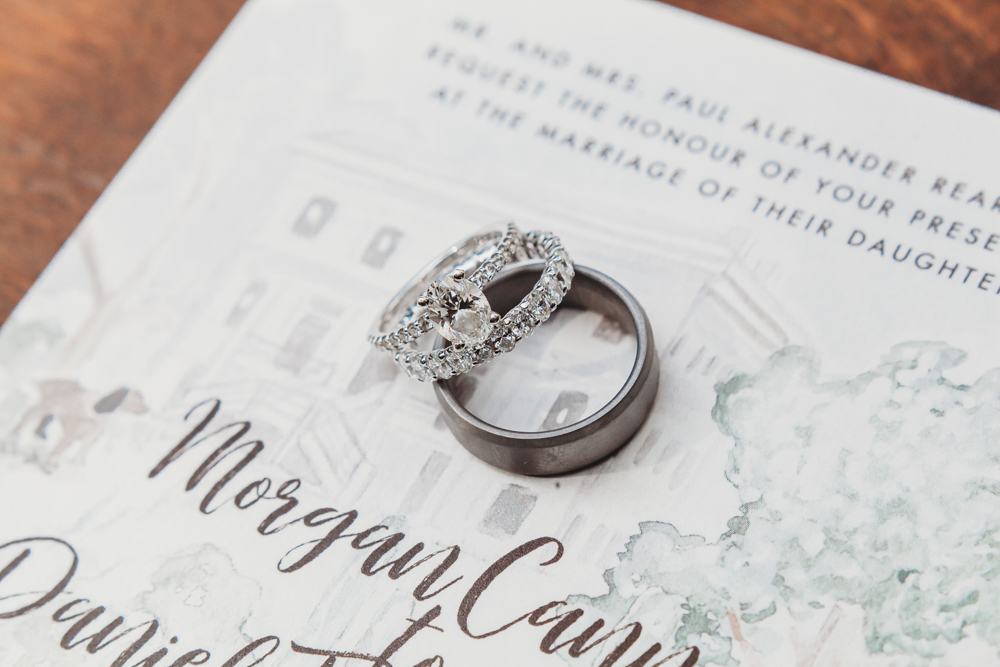 Diamond engagement ring and groom’s band resting on painted wedding invitation with soft watercolor florals.