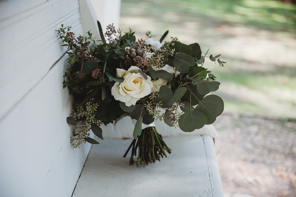 Bridal bouquet of white roses, eucalyptus, and greenery resting on whitewashed porch bench.