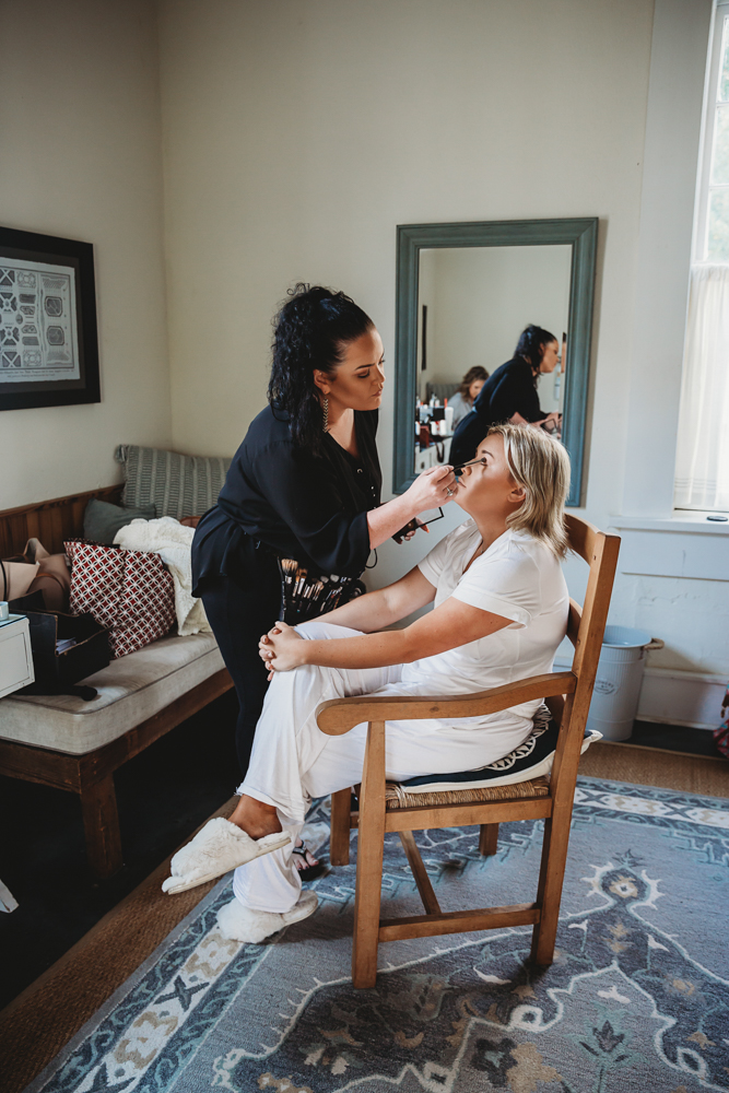 Bride getting makeup done by artist in cozy prep suite with patterned rug and morning light.