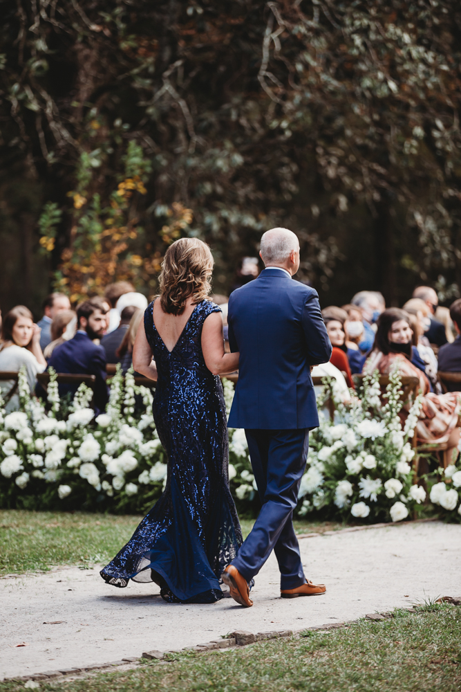 Bride’s parents walk down the floral-lined ceremony aisle, her mother in a navy sequined gown, captured at Wavering Place by Reflection Images by Tracy Rowell