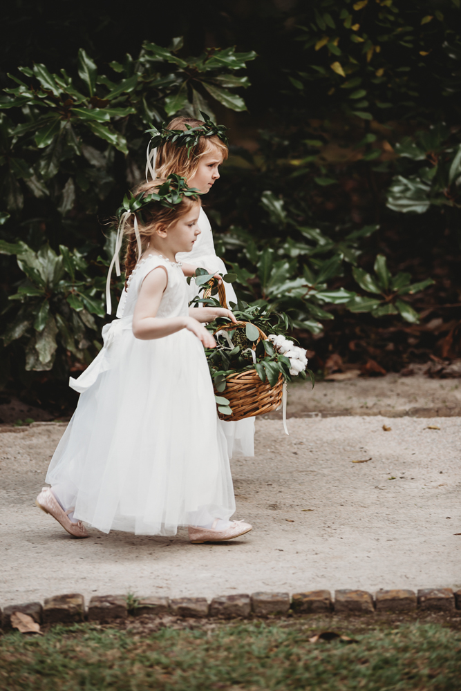 Side view of two flower girls walking the path with blush shoes, greenery crowns, and petal-filled baskets at Wavering Place