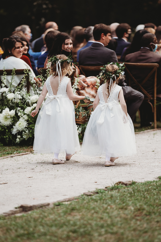 Flower girls in white tulle dresses and greenery crowns walk down the outdoor aisle with flower baskets at this SC wedding ceremony