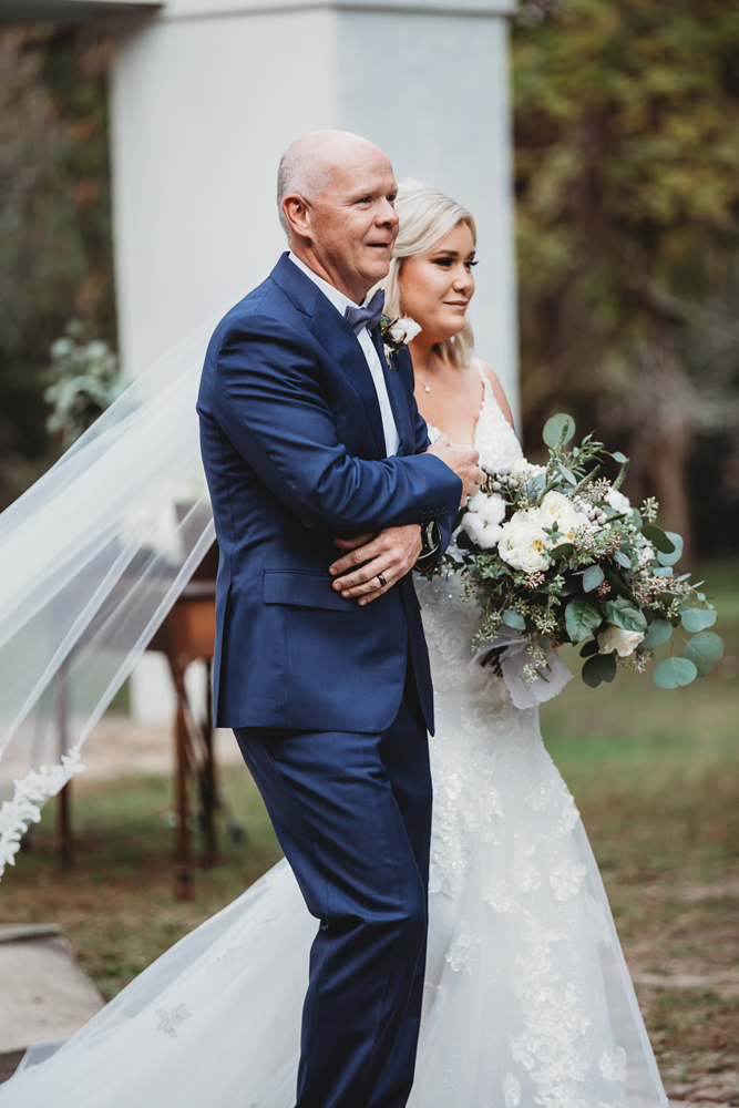 The bride walks arm in arm with her father, holding a bouquet of white blooms with cotton, her lace veil floating behind, photographed by Tracy Rowell wedding photography