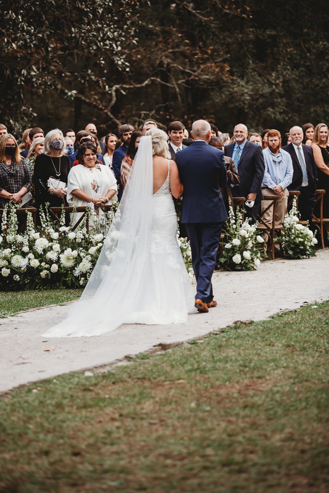 Bride and father walk toward the floral ceremony arbor while guests look on with smiles at Wavering Place, captured by SC wedding photographer Tracy Rowell