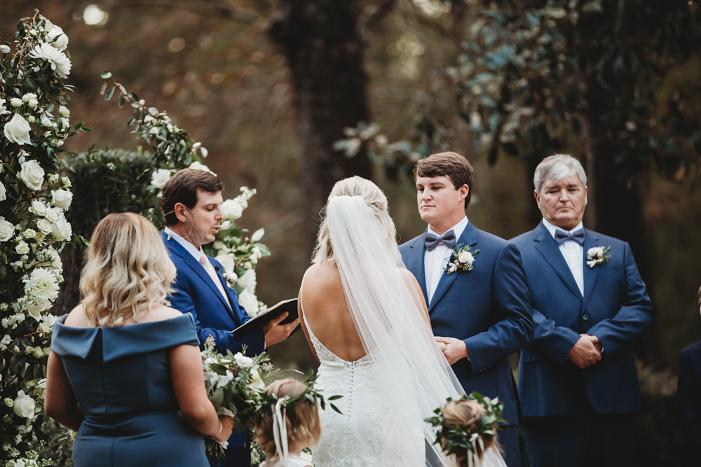 Bride and groom at the altar with blue-suited groomsmen and a lush white floral arch, taken by Reflection Images by Tracy Rowell