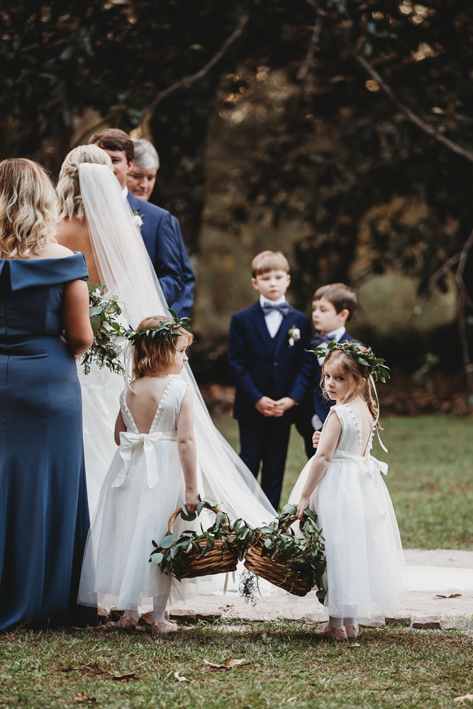 Flower girls holding greenery-filled baskets stand near young boys and bridesmaids during the ceremony at this elegant South Carolina wedding