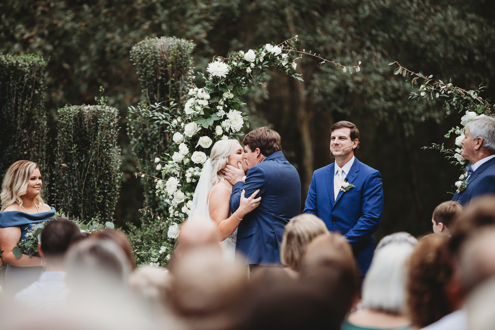 Bride and groom share their first kiss under a white rose floral arch, captured by Tracy Rowell wedding photography at Wavering Place in Eastover SC