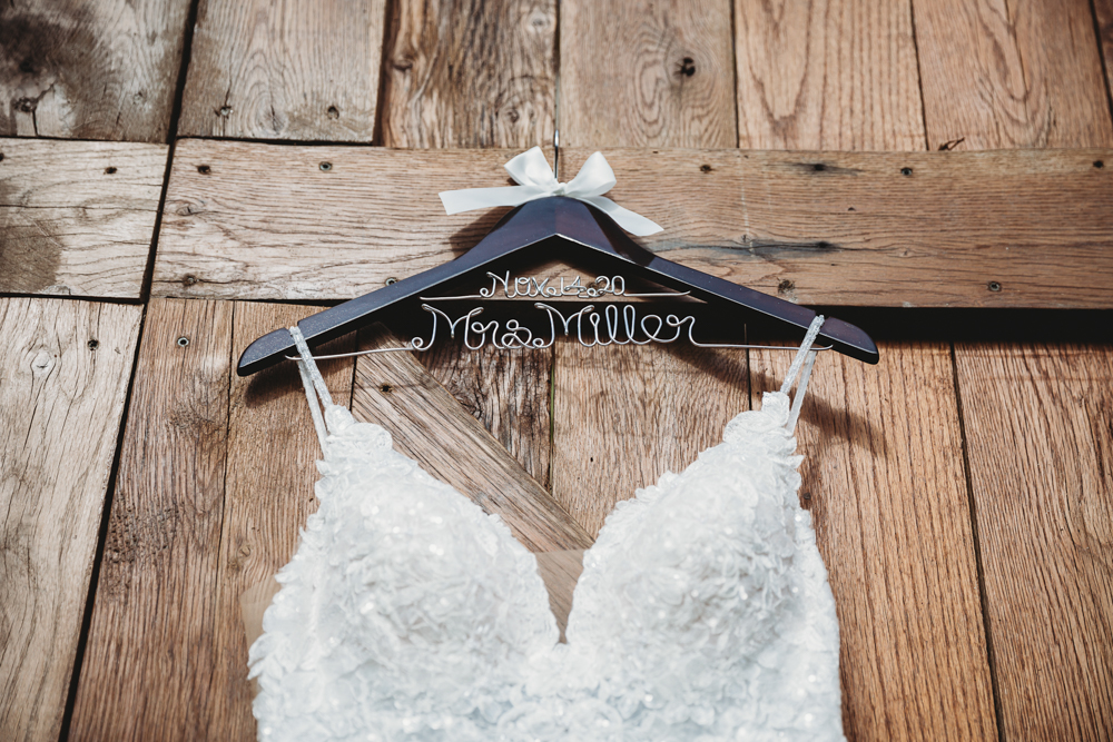 Lace bridal gown on custom hanger with 'Mrs. Miller' in wire and wedding date, styled against rustic wood paneling.