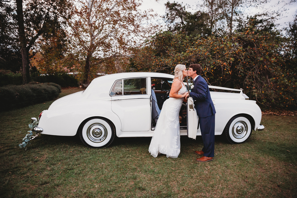 Bride and groom share a kiss beside a white vintage Rolls Royce with fall foliage behind at Wavering Place, photographed by Reflection Images by Tracy Rowell in Florence SC.