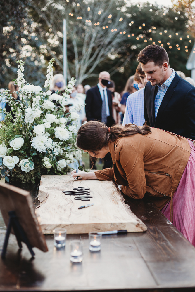 A guest signs a unique wooden guestbook surrounded by candlelight and a floral display of white roses at Wavering Place, photographed by Reflection Images by Tracy Rowell in Florence SC.