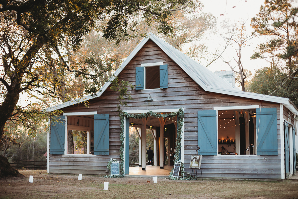 Wide view of the rustic blue-shuttered reception cottage at Wavering Place with framed bridal portraits and greenery-lined entrance, photographed by Reflection Images by Tracy Rowell in Florence SC.