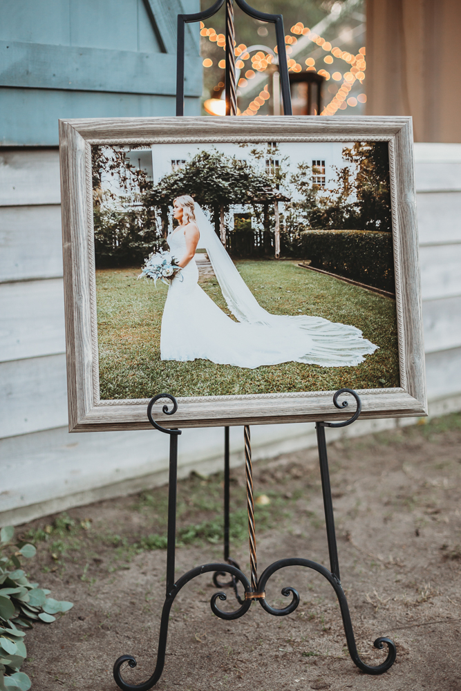 Framed full-body bridal portrait on easel outside Wavering Place reception area with twinkle lights and romantic garden backdrop, by Reflection Images by Tracy Rowell in Florence SC.