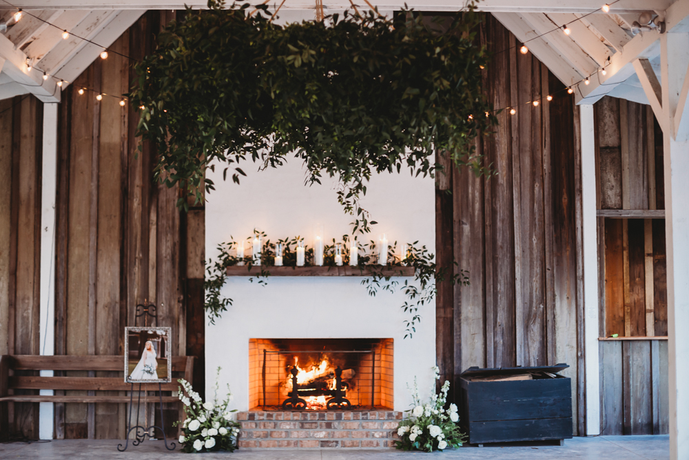 Cozy outdoor fireplace at Wavering Place decorated with white florals, greenery above, and framed bridal portrait nearby, photographed by Reflection Images by Tracy Rowell in Florence SC.