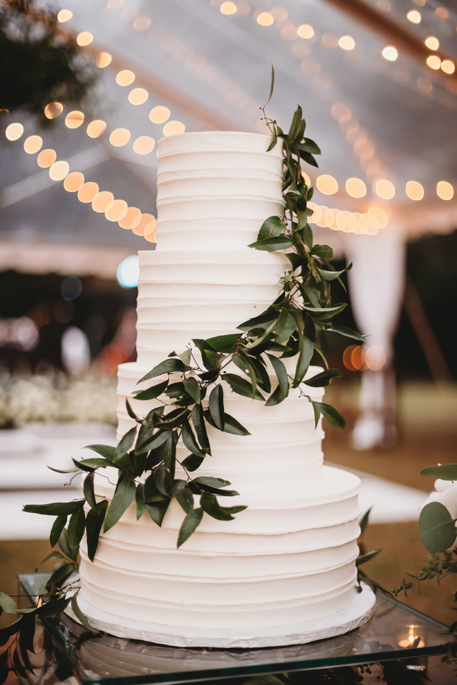Smooth white tiered wedding cake wrapped with cascading greenery, created by Cakes by Jeanie, set beneath string lights at Wavering Place, photographed by Reflection Images by Tracy Rowell in Florence SC.
