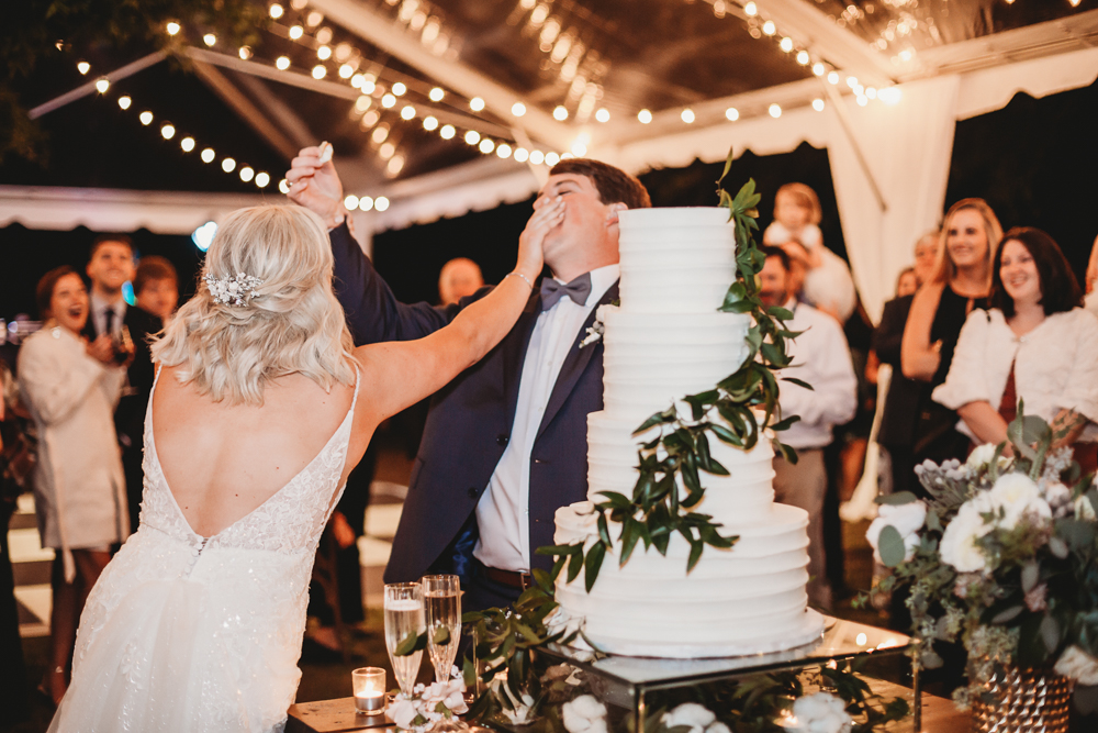 Bride playfully smashes cake into the groom’s face as guests laugh during the fun reception at Wavering Place, by Reflection Images by Tracy Rowell in Florence SC.