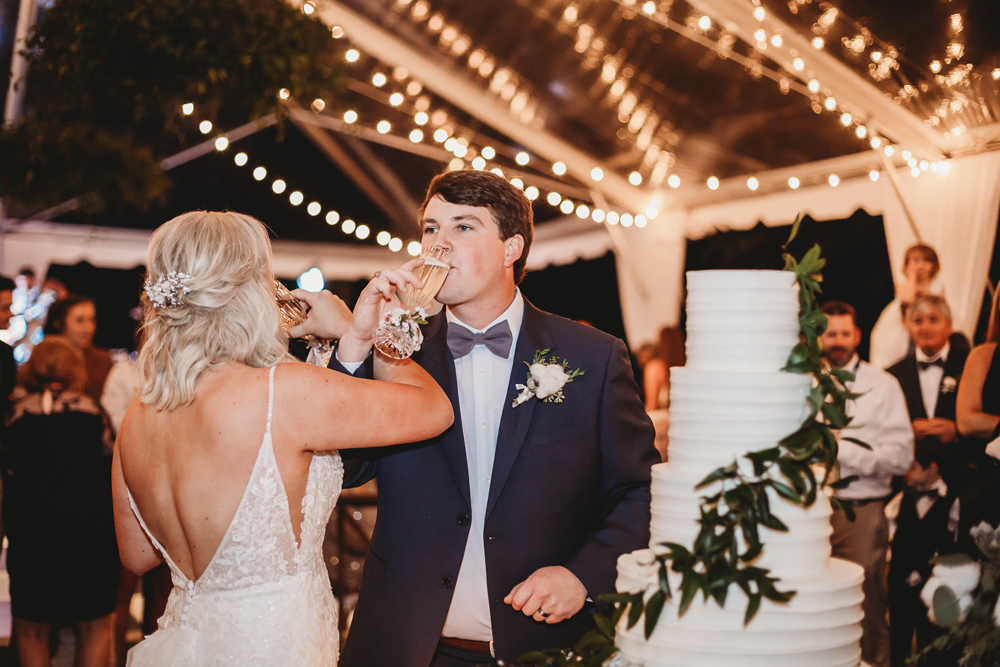 Bride and groom clink champagne flutes by their tiered white cake wrapped in greenery at Wavering Place, photographed by Reflection Images by Tracy Rowell in Florence SC.