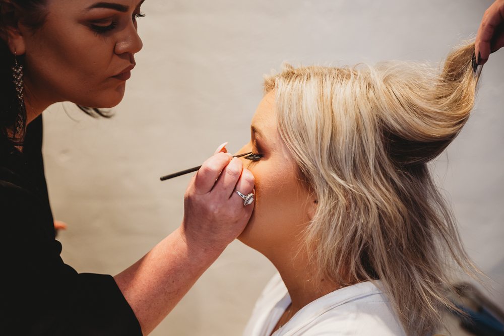 Close-up of bride having eyeliner applied by makeup artist, showing detailed eye makeup and blonde hair.
