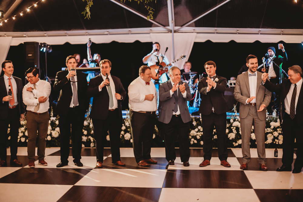 Groomsmen and family line up with drinks in hand for a celebratory toast on the dance floor at Wavering Place, with live band in the background, by Reflection Images by Tracy Rowell in Florence SC.