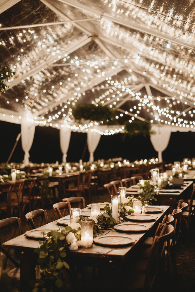 Long tables under a clear tent at Wavering Place, beautifully decorated with candlelight, greenery, and string lights overhead during the reception.