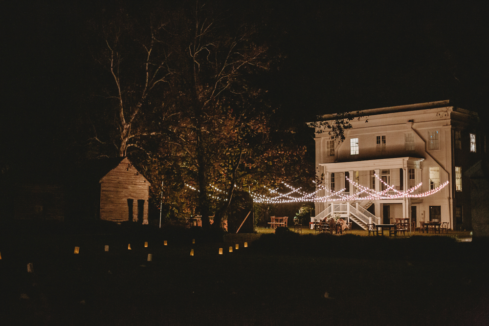 The Wavering Place house glowing at night with string lights and lantern-lit pathways, setting a romantic atmosphere for the celebration.