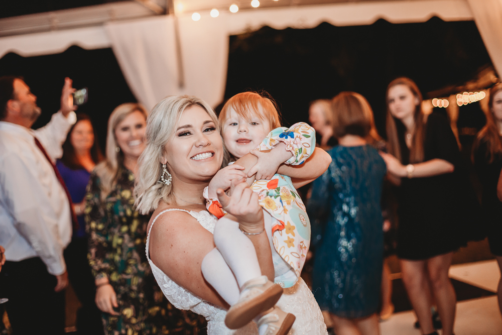 Bride Morgan holding a smiling toddler in a colorful dress while dancing joyfully during the reception at Wavering Place.