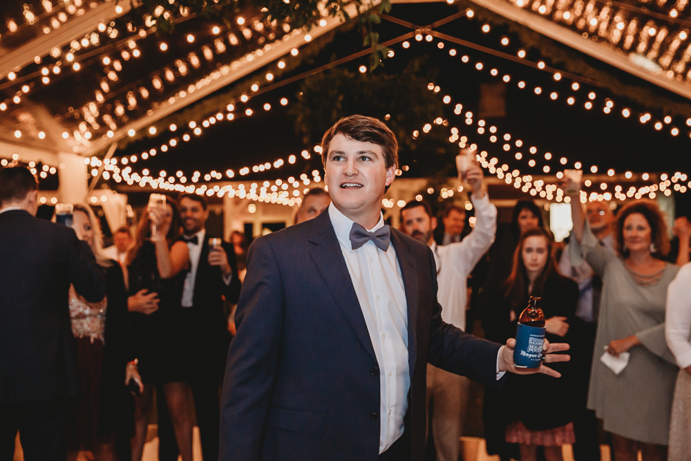 Groom Daniel raising a bottle in a toast surrounded by cheering guests and sparkling string lights at the Wavering Place reception.