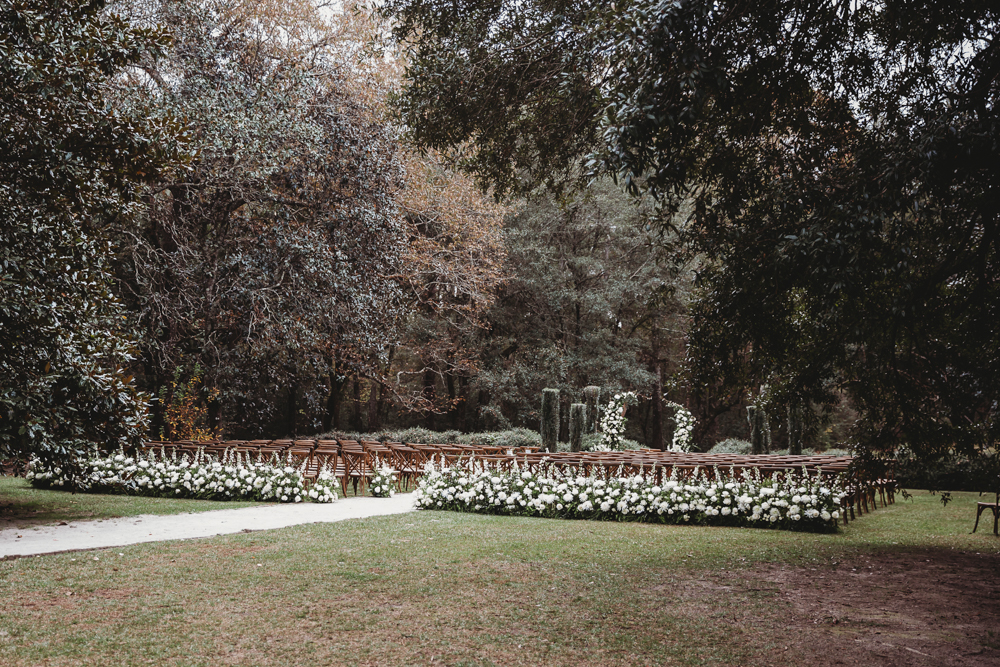 Wide view of outdoor ceremony setup with white florals, wood chairs, and forest backdrop at Wavering Place.