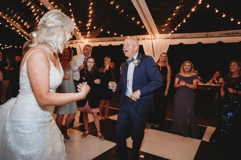 The bride and her father share a joyful dance together under a canopy of glowing string lights, surrounded by smiling guests watching and cheering them on.