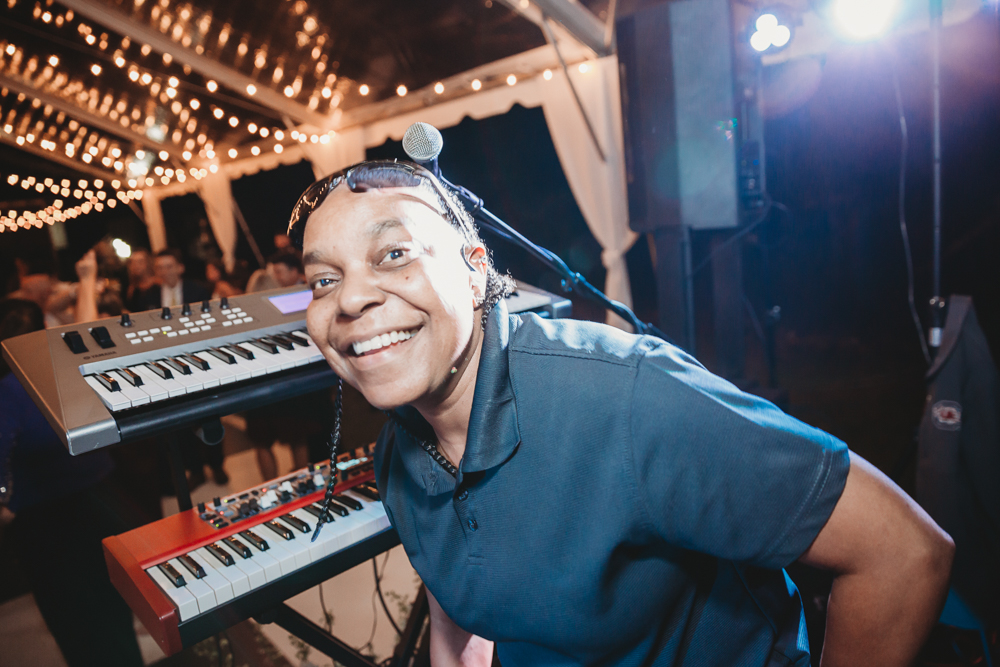 A close-up of a smiling band member with a headset mic in front of two keyboards, lit by blue stage lights and surrounded by wedding guests in the background.