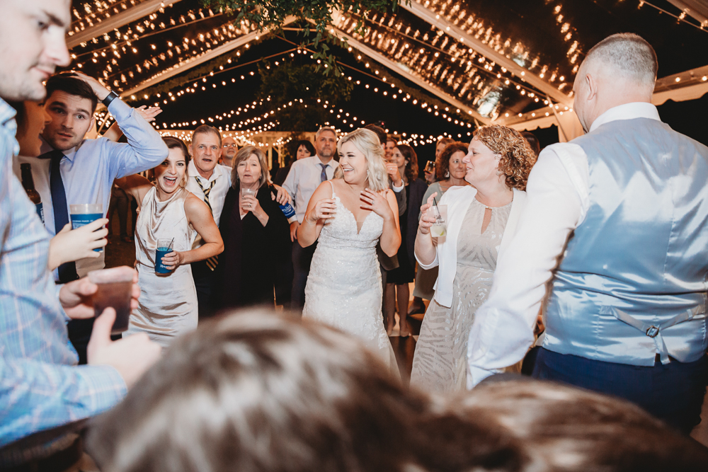 The bride is surrounded by friends and family on the dance floor, all holding drinks and celebrating together with big smiles.