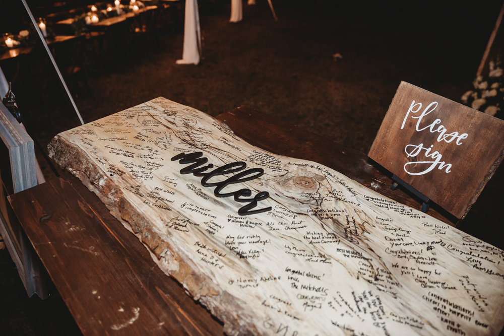A rustic wooden guest sign-in slab engraved with “Miller,” covered in handwritten messages, sits beside a wooden sign that says “Please sign.”