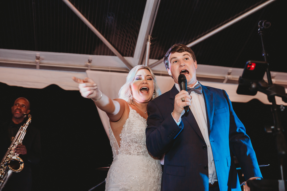 The bride and groom joyfully sing into microphones on stage, pointing and engaging with the crowd while a saxophonist plays behind them.
