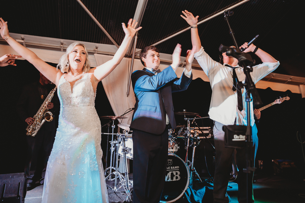 The bride and groom raise their arms with the band, celebrating on stage after a performance, in front of the drum kit with the band's logo “MB.”