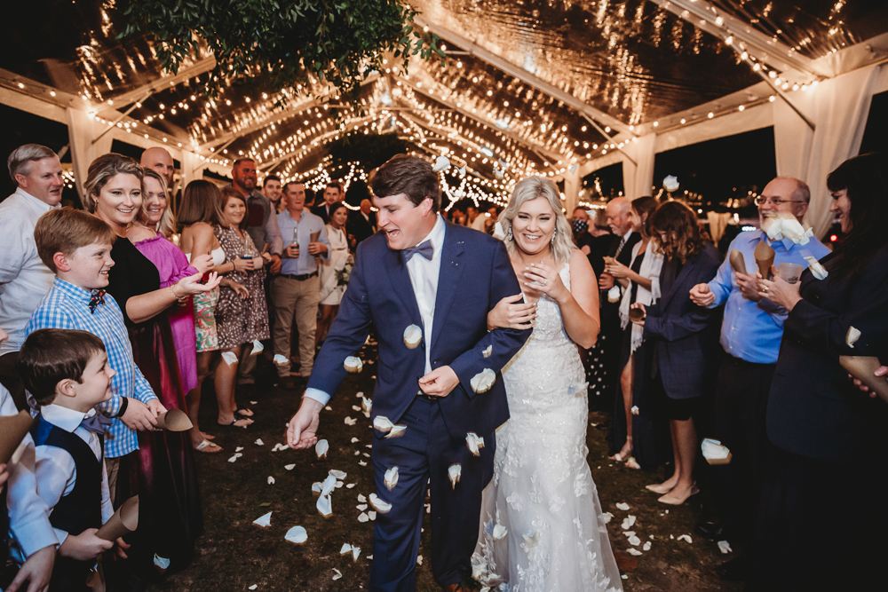 The newlyweds make a festive exit through a petal toss, laughing as guests cheer and shower them with flower petals under the tent lights.