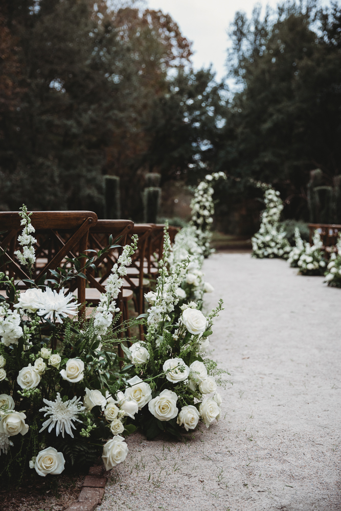 Elegant white roses and snapdragons lining the aisle with wooden chairs and trees in the background.
