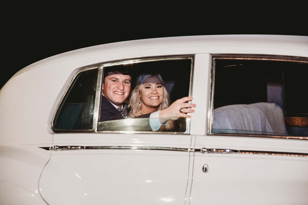 The bride and groom smile and wave from the backseat of a classic white car, ready to depart, beaming with happiness.