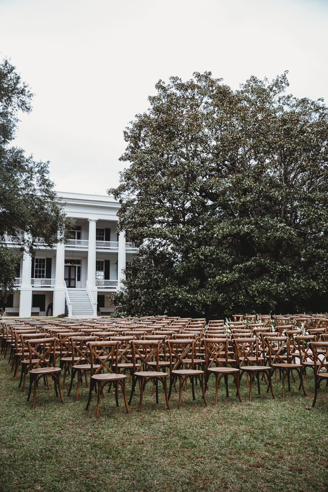 Rows of wooden cross-back chairs set up on the lawn in front of the grand white-columned house.